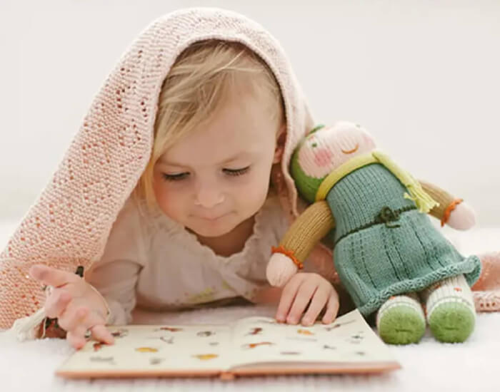 Young child with blanket draped on head reading book next to stuffed doll