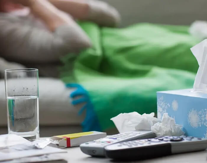 A sick person lying on a sofa and covered with a blanket. In the foreground discarded tissues rest on top of television remote controls.