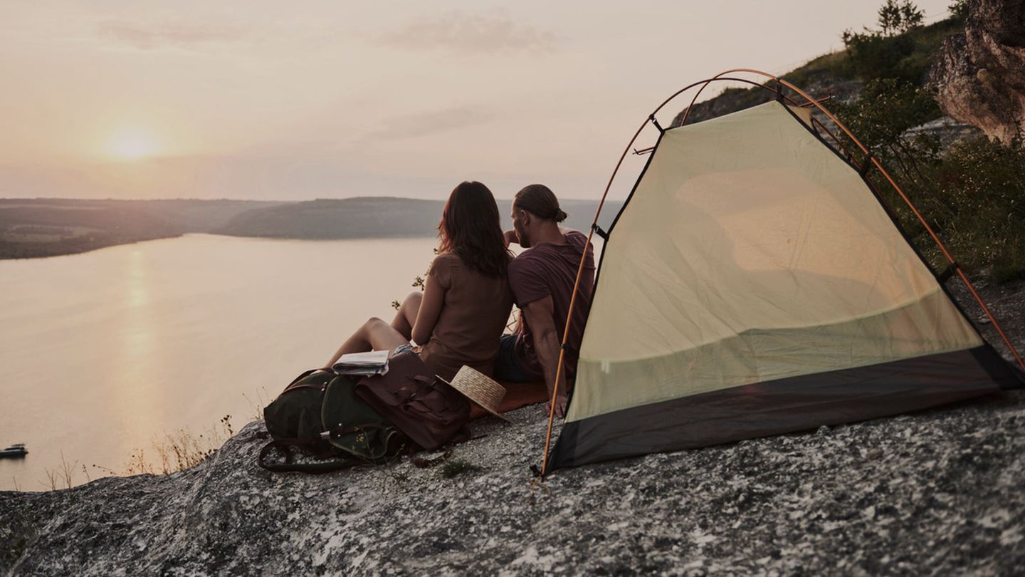 Young couple sit in front of their tent, side-by-side, leaning towards one another while watching the sun set above a body of water.