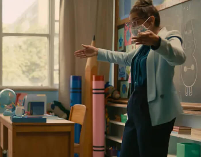 A teacher wearing a cloth face mask standing at the front of a classroom containing mask-wearing young children at widely separated desks. The chalkboard in the background features cartoons of germs.