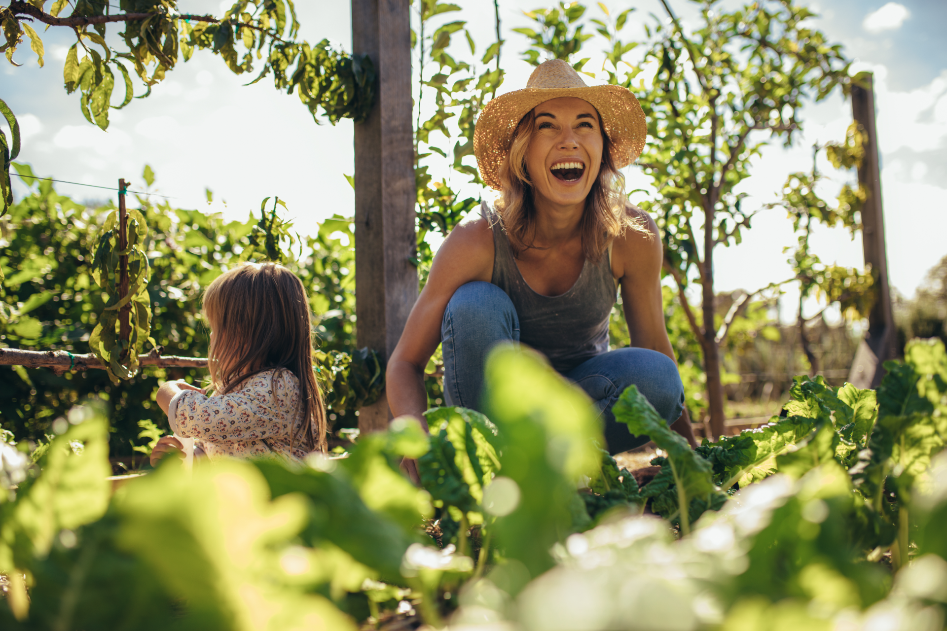 A woman wearing a hat and smiling sits in a garden, while a young girl nearby looks at plants.