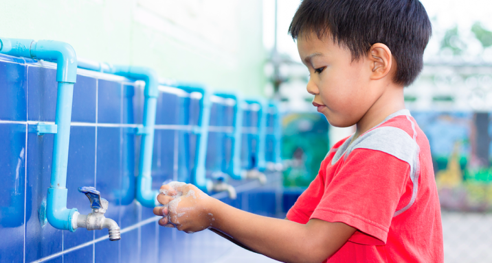 A young boy in a red shirt washes his hands at a row of blue tiled sinks with blue pipes, concentrating on cleaning his hands thoroughly.