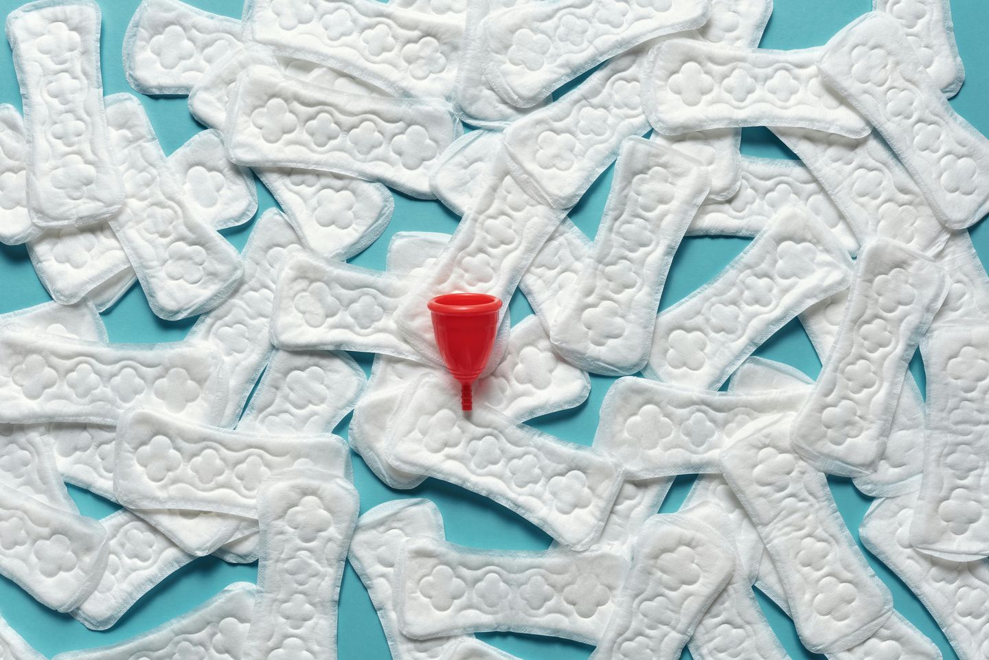 Absorventes femininos descartáveis espalhados sobre fundo azul com coletor menstrual vermelho ao centro.