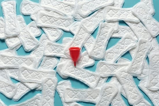 Absorventes femininos descartáveis espalhados sobre fundo azul com coletor menstrual vermelho ao centro.