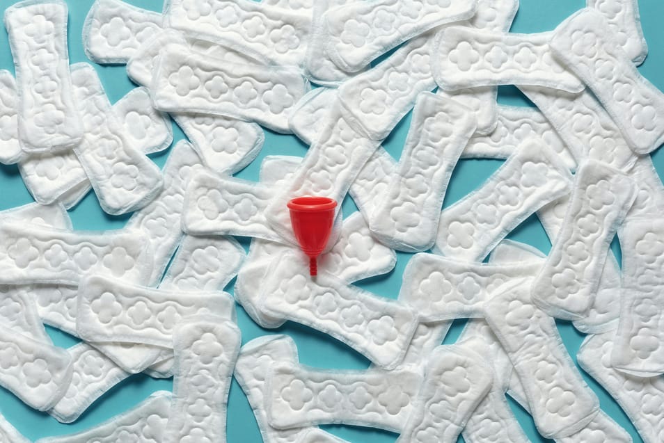 Absorventes femininos descartáveis espalhados sobre fundo azul com coletor menstrual vermelho ao centro.