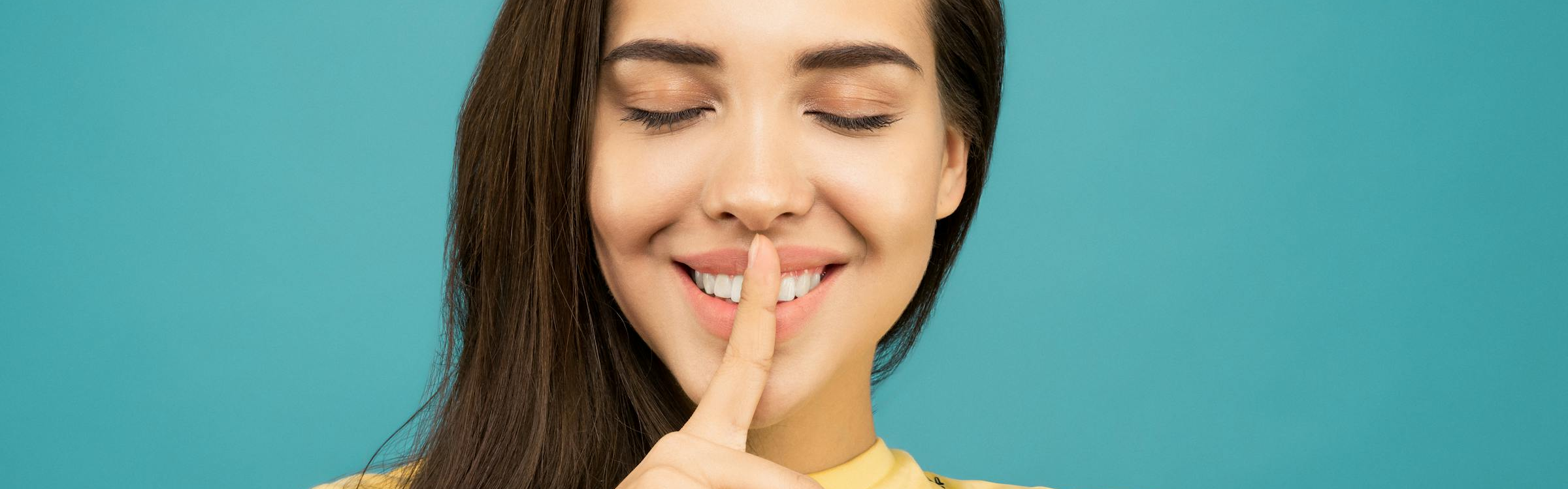 Close-up of a person with eyes closed, holding one finger vertically against their lips in a “quiet” or “shh” gesture, against a teal background