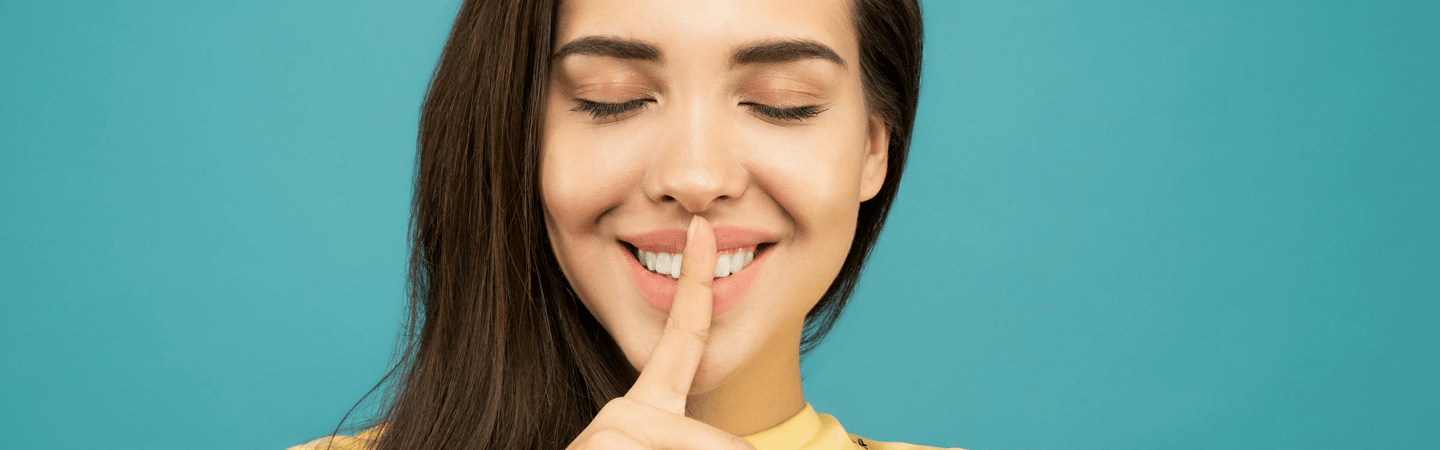 Close-up of a person with eyes closed, holding one finger vertically against their lips in a “quiet” or “shh” gesture, against a teal background