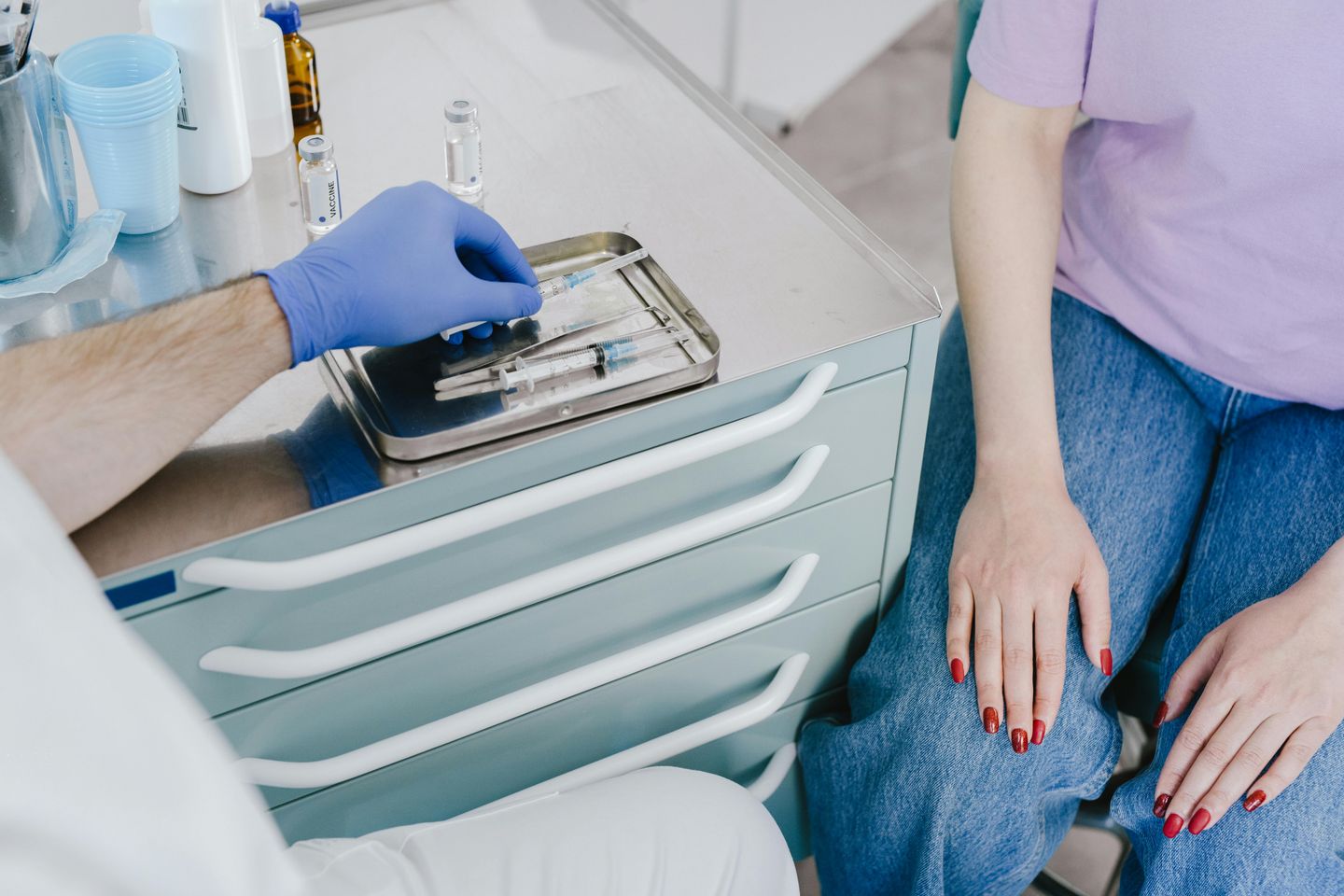 Profissional da saúde preparando seringa com vacina enquanto paciente aguarda sentada, com as mãos visíveis, na sala de atendimento.