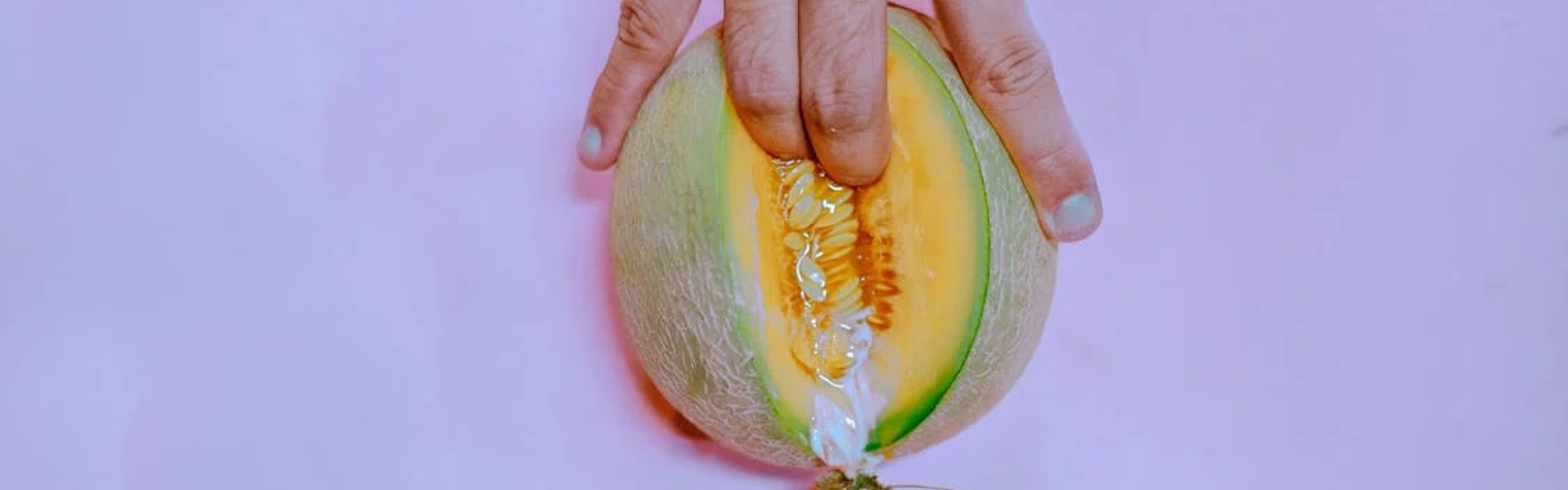 A close-up image of a hand pressing into a halved melon, with visible seeds and juice, set against a soft pink background.