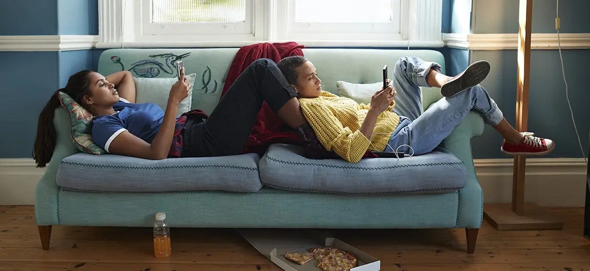 Two teenagers relaxing on a sofa and playing on their phones. They are lying sideways on the cushions of the sofa and are resting on each other and the arms of the chair.