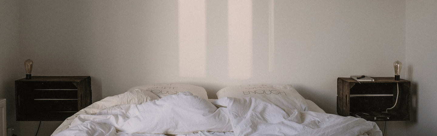 Unmade bed with white bedding between two wooden nightstands, each holding a small lamp, in a softly lit minimalist bedroom.