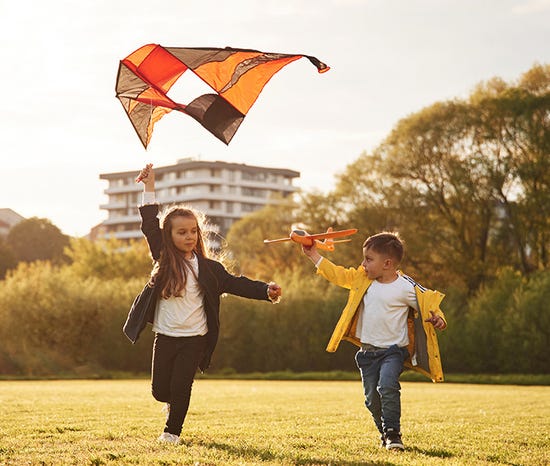 Two children playing in a grassy field with a kite and toy airplane. Trees and a building in the background.