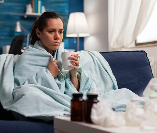 Person wrapped in a blanket holding a mug on a couch, with medicine bottles and tissues on a table.