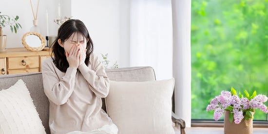 A person sitting on a sofa covering their face, with a wooden cabinet behind and green foliage visible through a window.