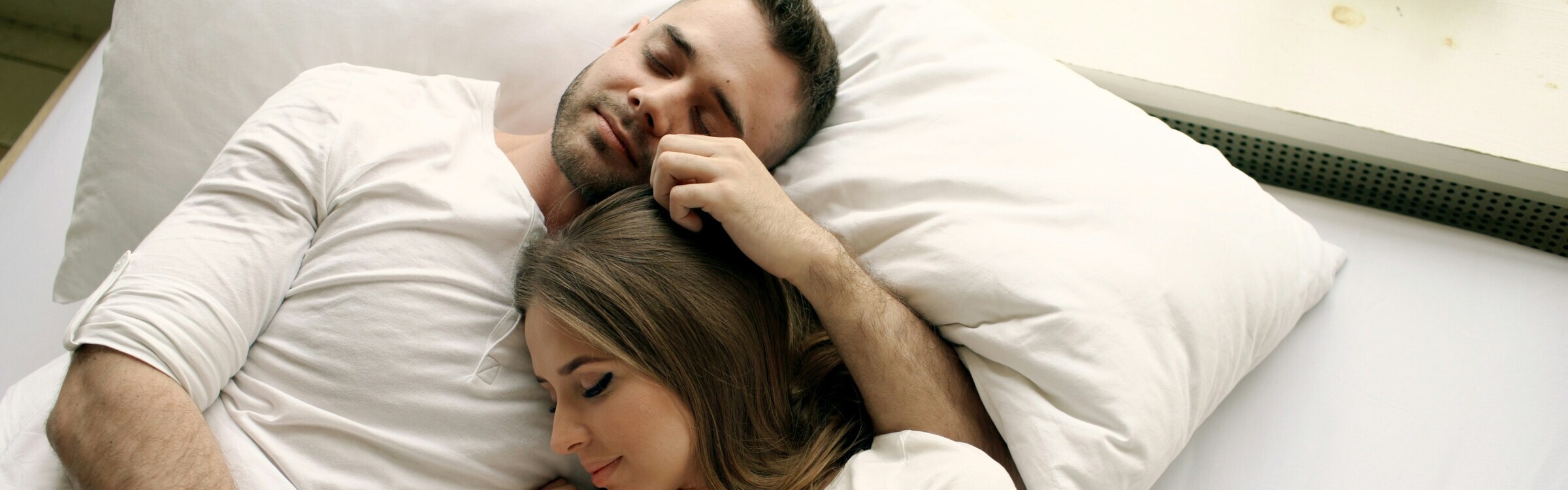 Two people lying on a bed with white bedding, one resting their head on the other’s chest while the other person gently places a hand on their head.