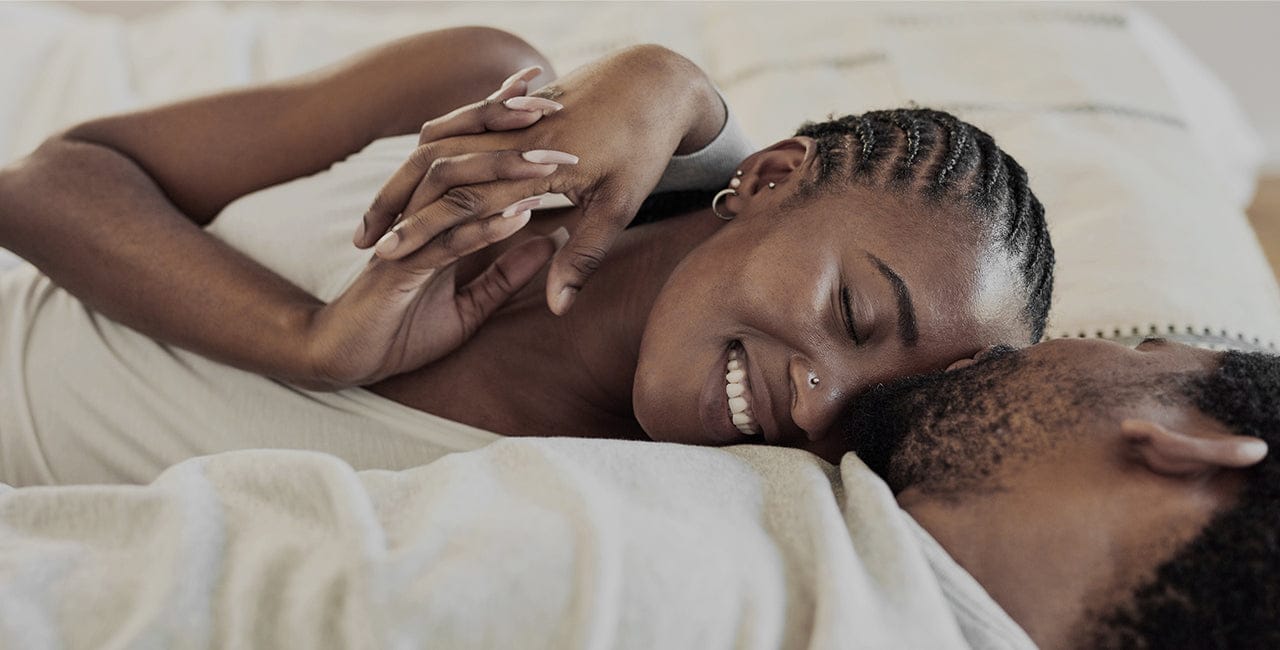 A couple enjoys a tender moment on a bed; the woman with braided hair smiles joyfully at the man whispering in her ear, in a softly lit bedroom.