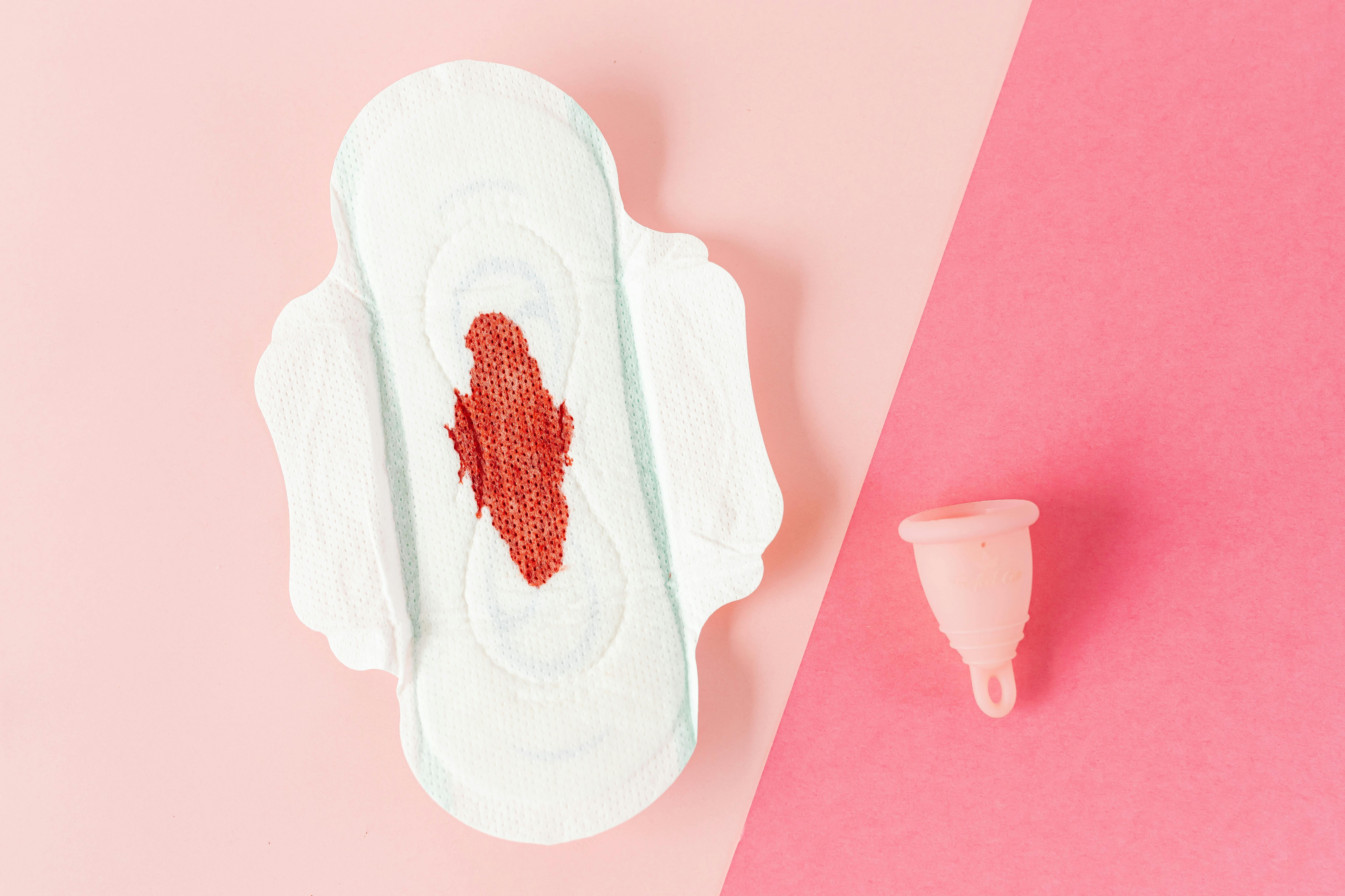 Absorvente feminino com mancha de sangue ao lado de coletor menstrual sobre fundo rosa.