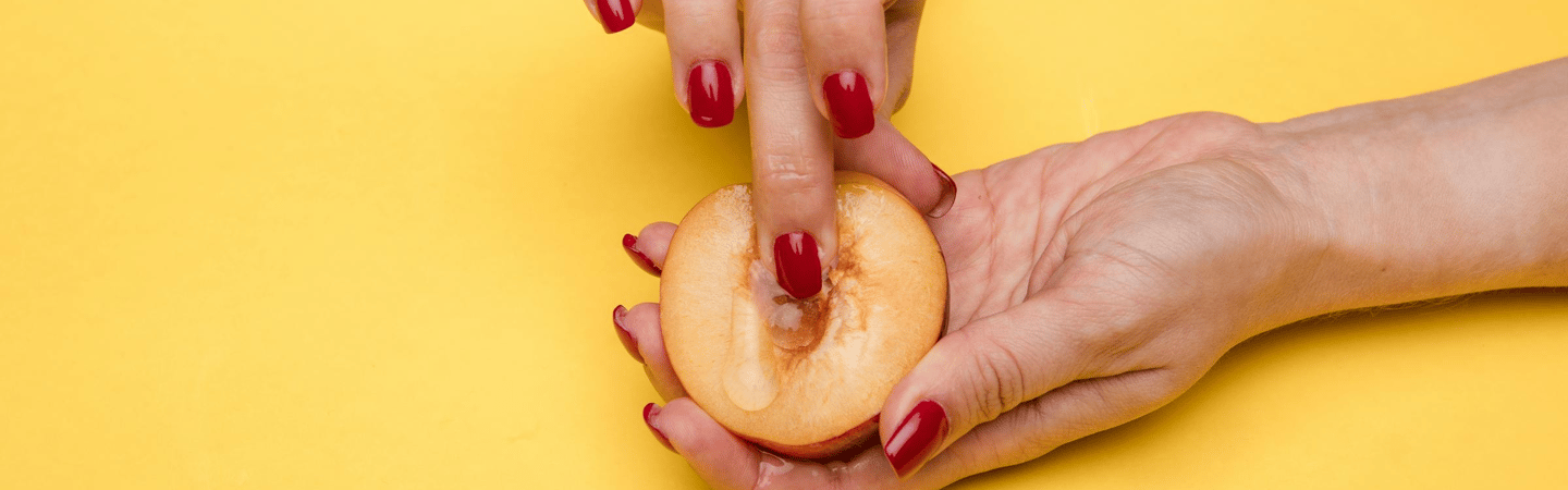 Close-up of hands with red nail polish holding a halved peach against a yellow background, with liquid on the fruit surface.