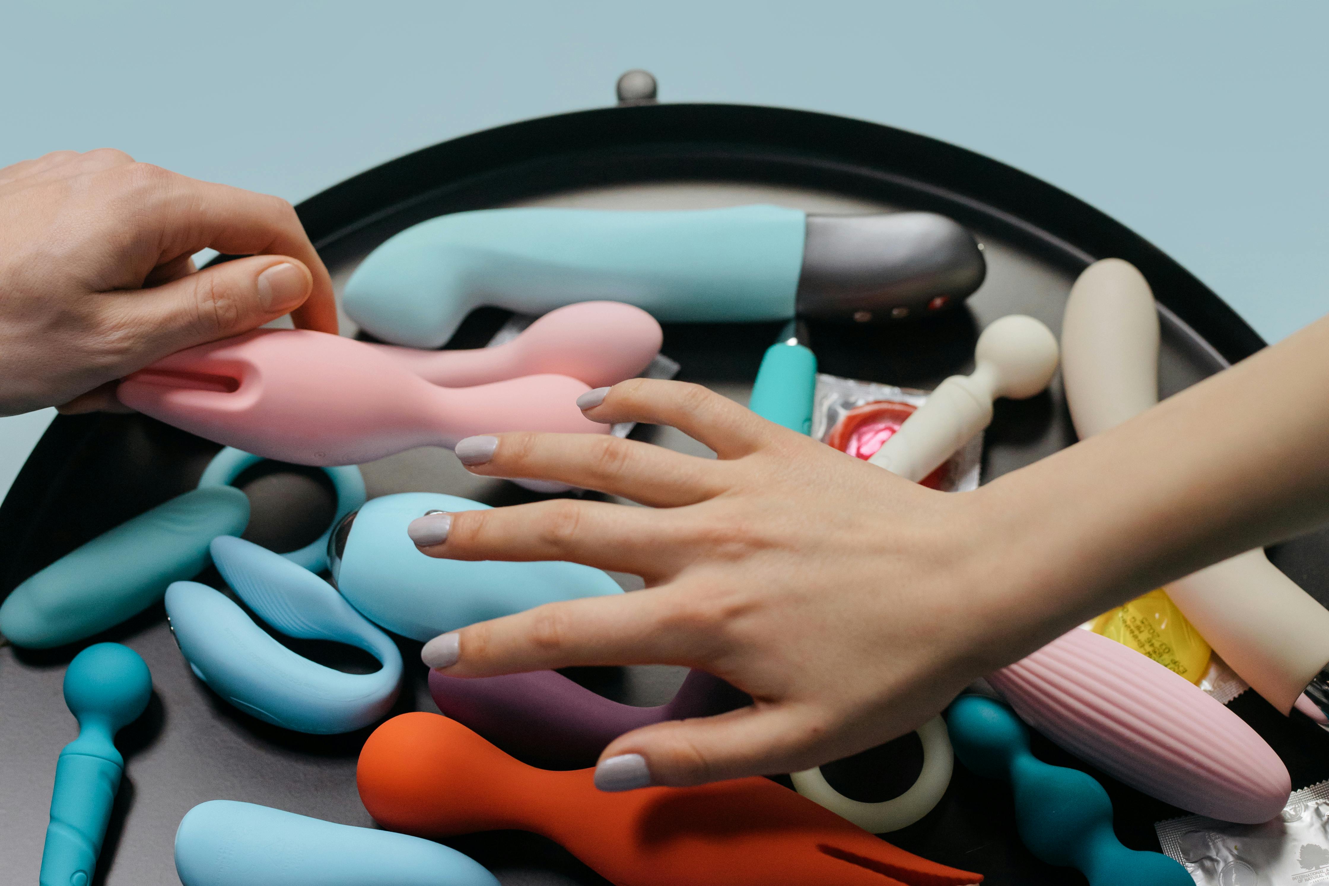 Mãos escolhendo vibrador entre diversos brinquedos íntimos coloridos sobre bandeja