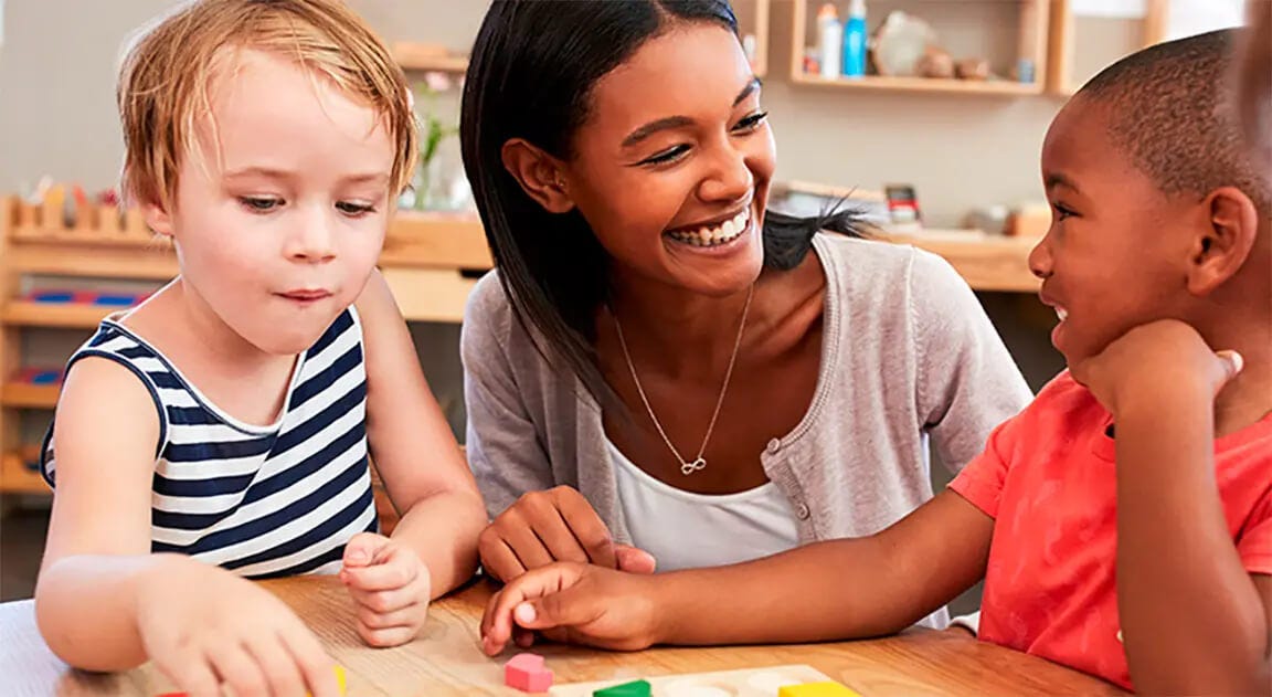 Teacher smiles at young children sitting at a classroom desk