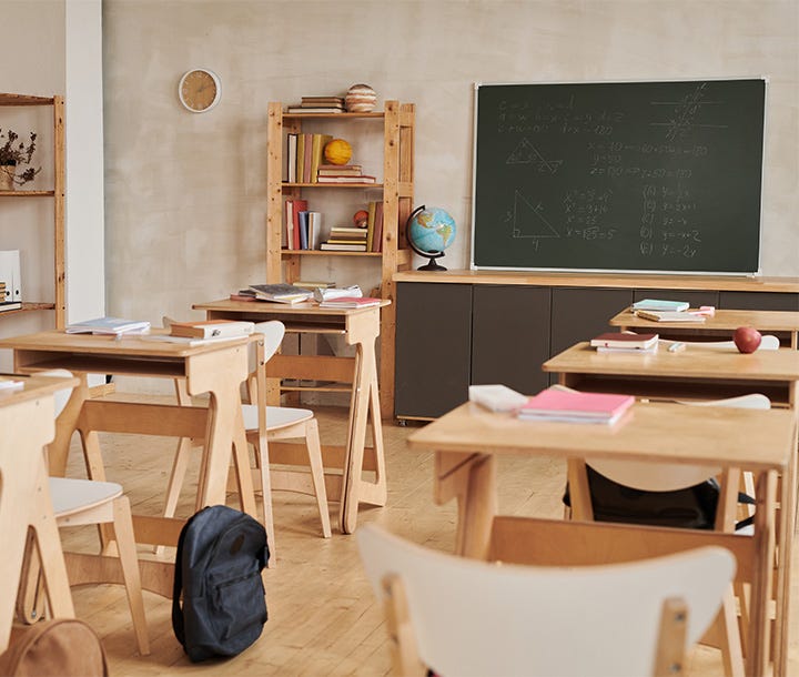 A classroom with wooden desks, chairs, a chalkboard with geometric drawings, and a shelf with books and a globe.