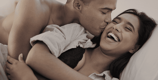 Un couple rit en se câlinant au lit, pendant que l’homme embrasse doucement la joue de la femme.