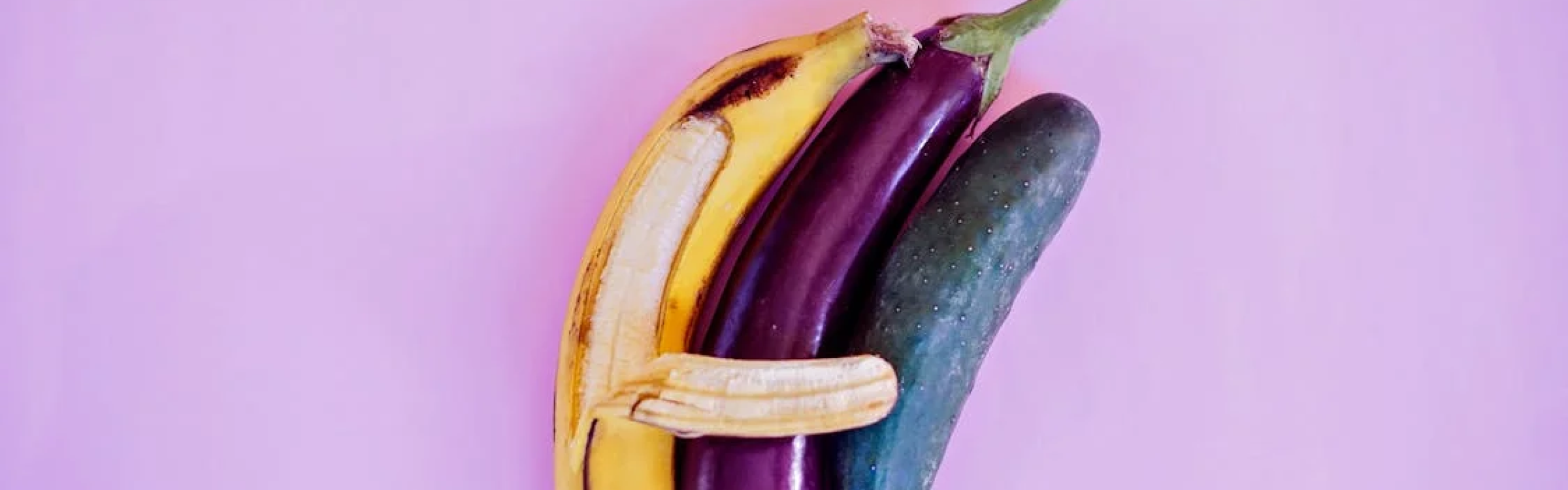 A peeled banana hugging an eggplant and a cucumber against a pink background.
