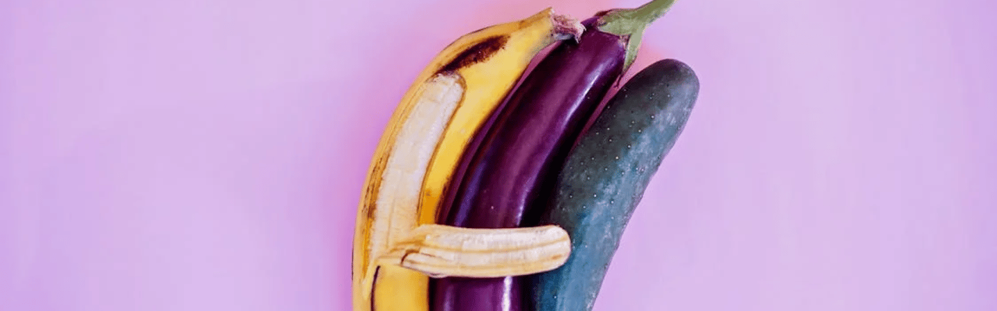 A peeled banana hugging an eggplant and a cucumber against a pink background.