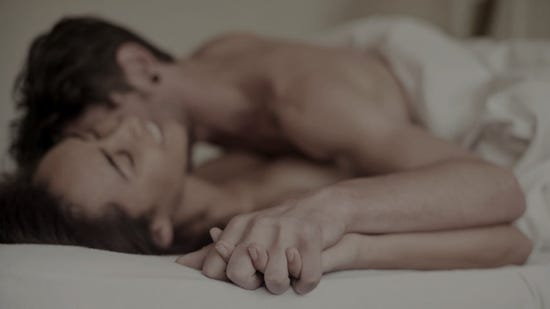 Man lying on top of a smiling woman in bed whispering his desires into her ear and holding her hand.