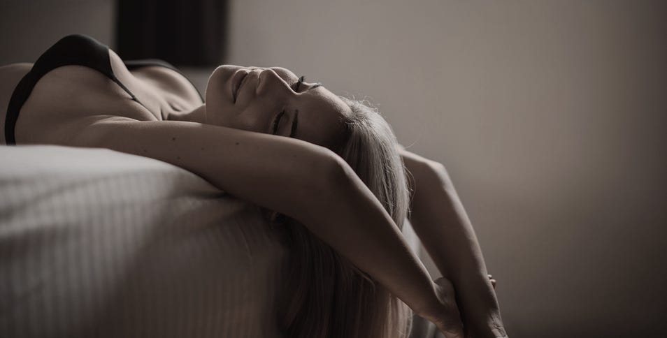 A woman with long hair reclines backward on a bed, her arms stretched above her head in a sensual pose filled with quiet pleasure.