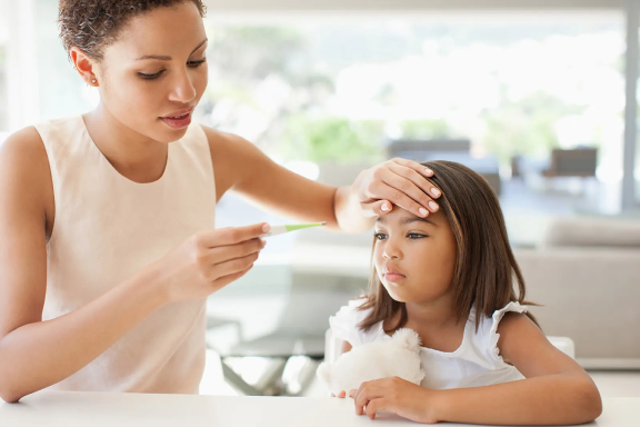 Adult holding a thermometer towards a child holding a stuffed toy at a table.