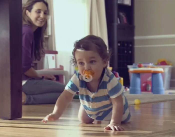 A parent kneeling on the floor and smiling at their baby, who is crawling away
