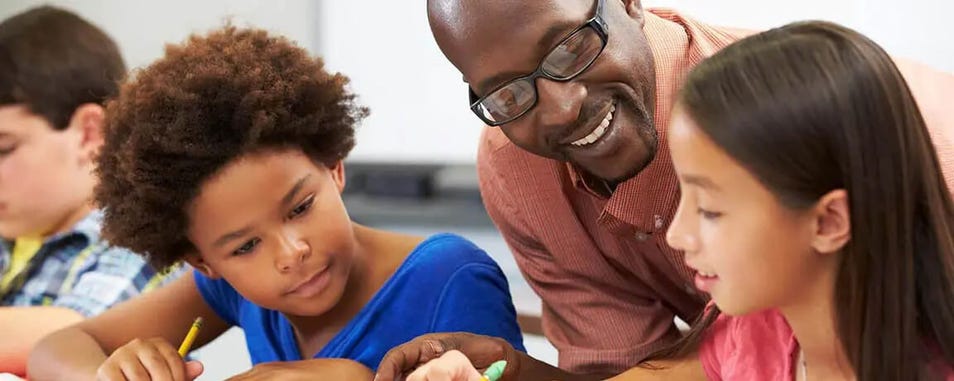 Teacher smiling while leaning over children who are writing in workbooks