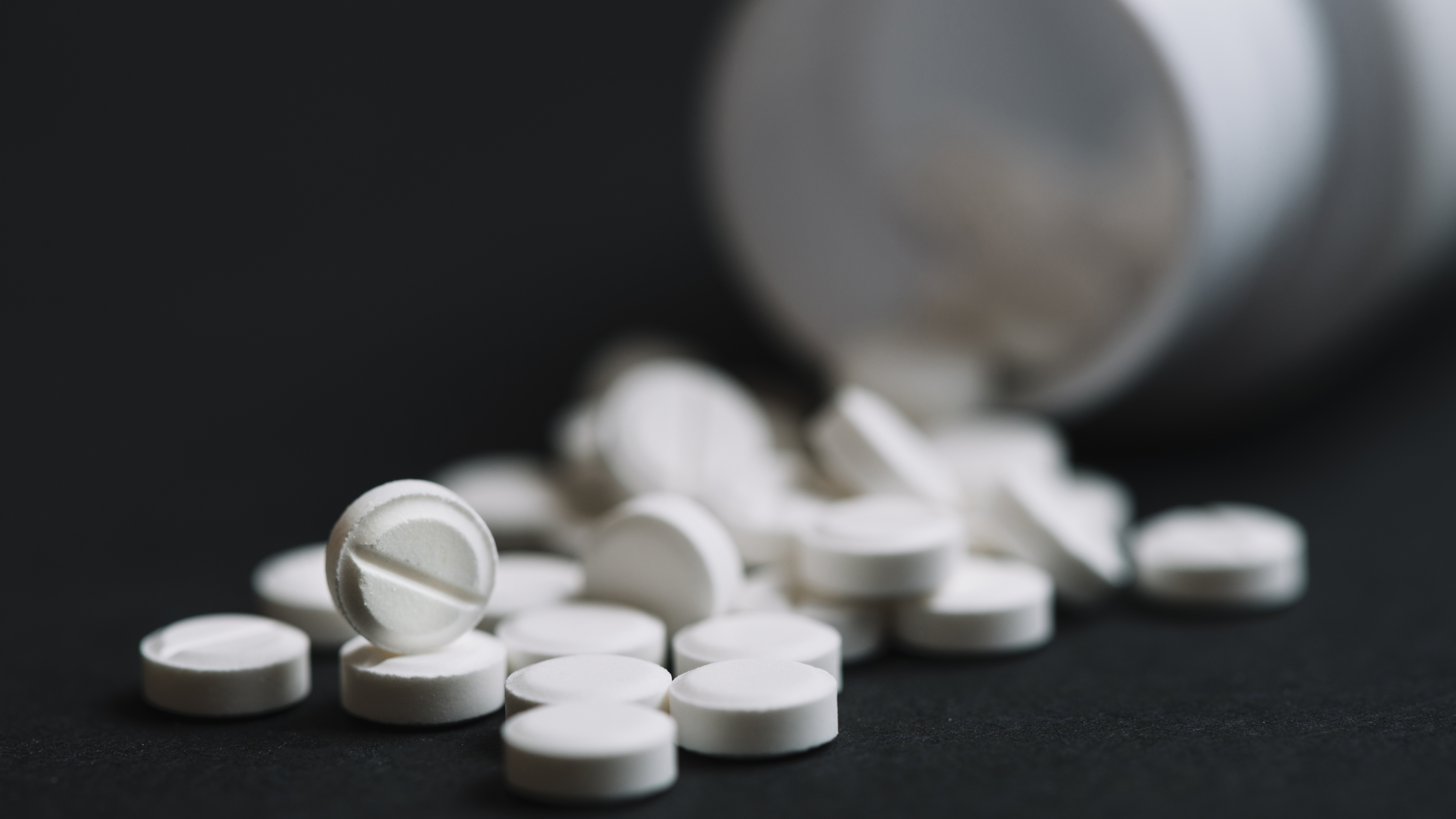 Comprimidos brancos espalhados ao lado de um frasco de medicamento em fundo escuro.