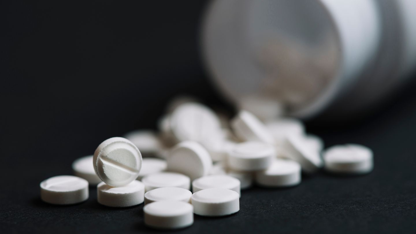 Comprimidos brancos espalhados ao lado de um frasco de medicamento em fundo escuro.