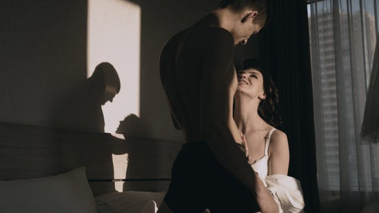 Woman in white kneeling before her partner as a shadow of their silhouette is casted on the bedroom wall.