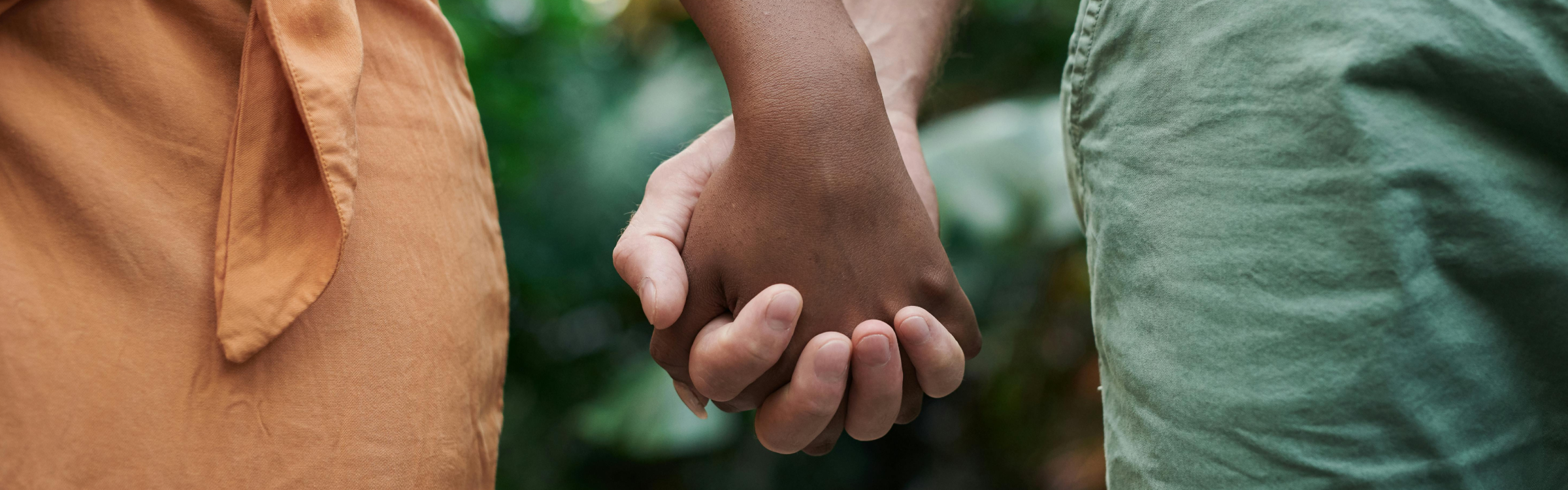 Close-up of two people holding hands, standing side by side outdoors with blurred greenery in the background, symbolizing connection and togetherness.