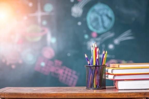 Colorful pencils in a holder next to stacked books on a desk, with a chalkboard background featuring drawings of atoms and molecules.