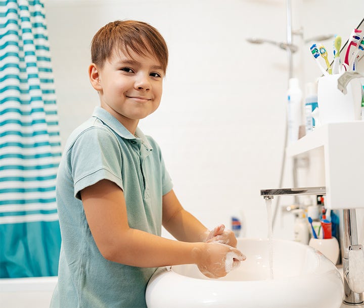 A boy washing his hands at a bathroom sink with a blue and white shower curtain in the background.