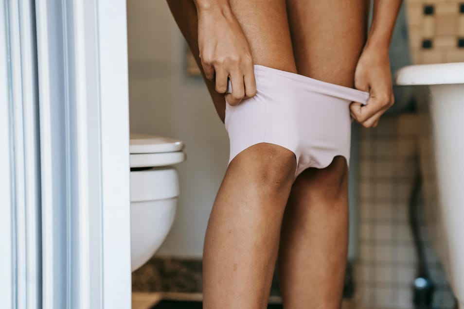 Pessoa abaixando a roupa íntima no banheiro, em frente ao vaso sanitário, em ambiente doméstico.