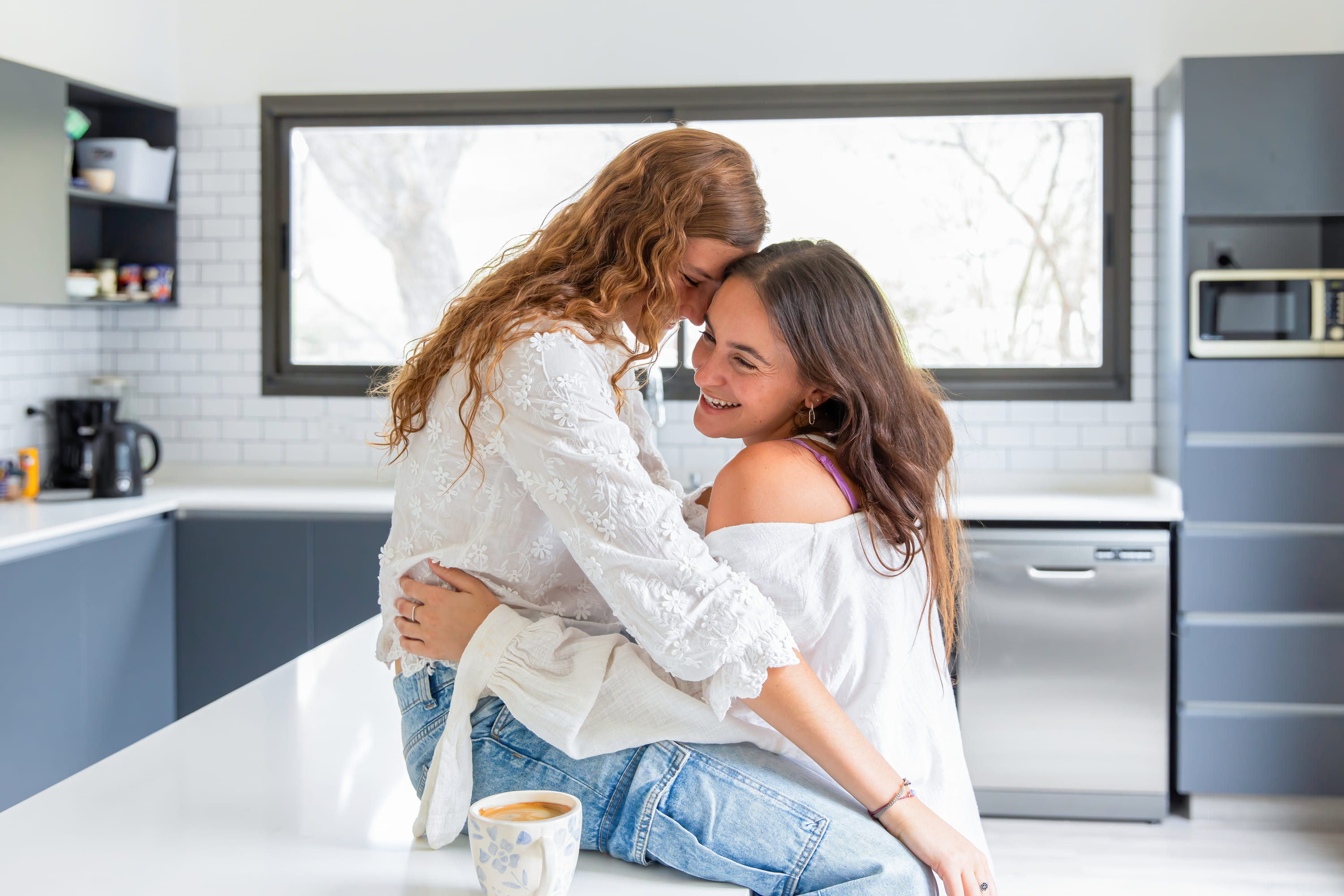 Una pareja de mujeres sentadas en la cocina diciendose secretos al oido