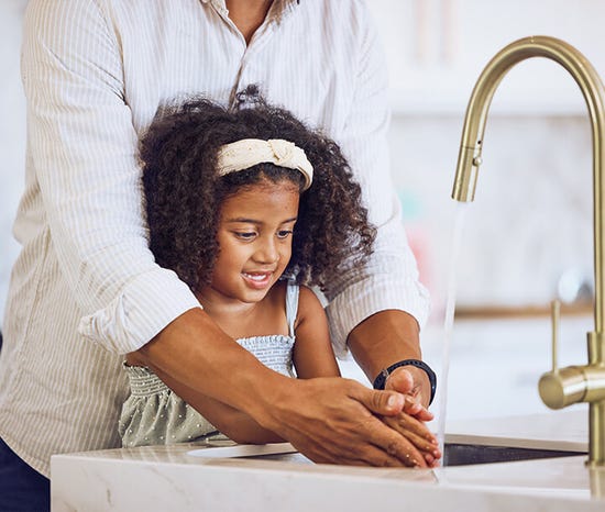 Adult helping a girl wash her hands at a kitchen sink.