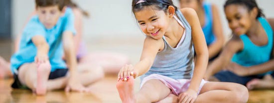 Children stretching on a wooden floor, with a smiling girl in a grey tank top reaching for her foot.