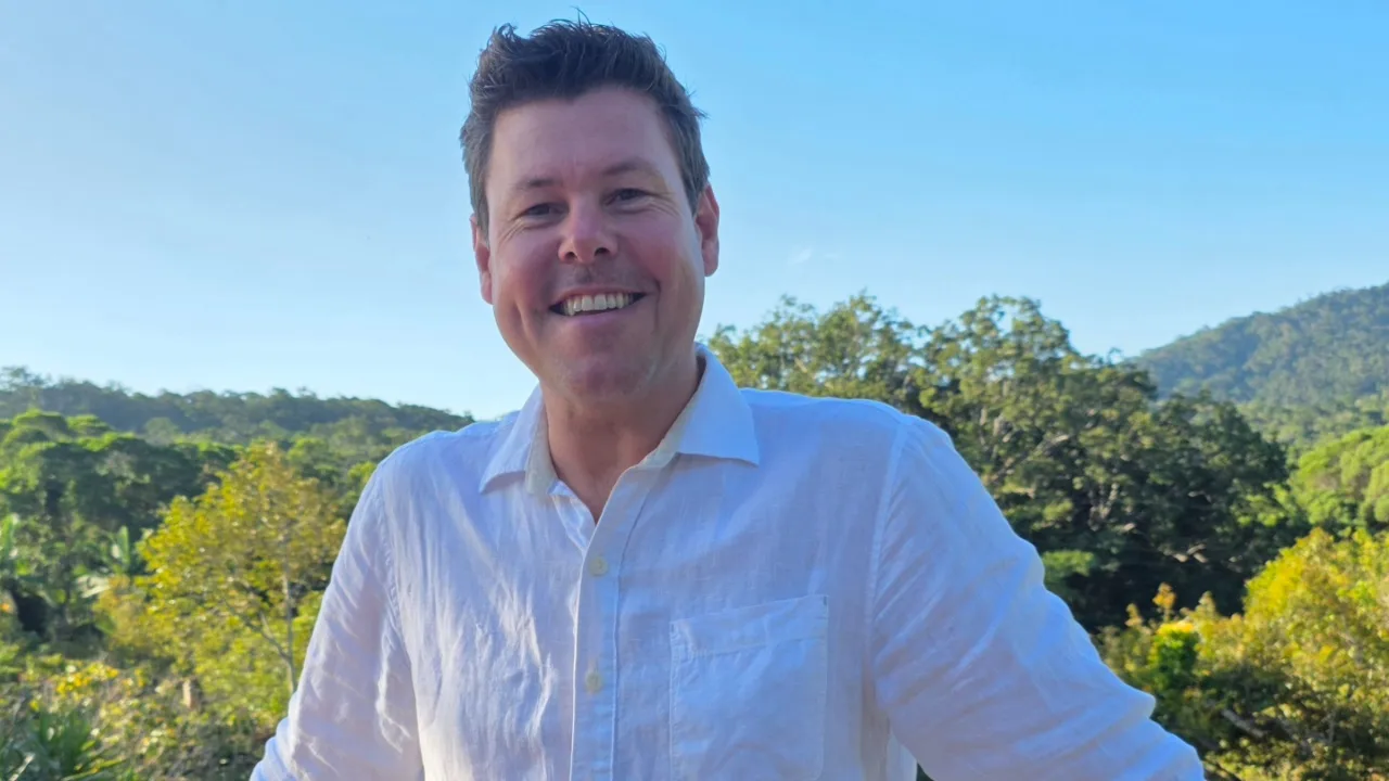 A smiling man with light brown hair wearing a white shirt stood before a canopy of trees