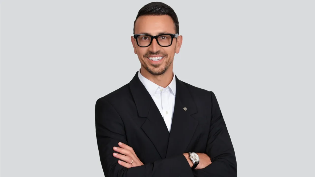 Um homem sorridente de cabelo preto, casaco de fato azul-marinho, camisa branca, de óculos, está de braços cruzados.