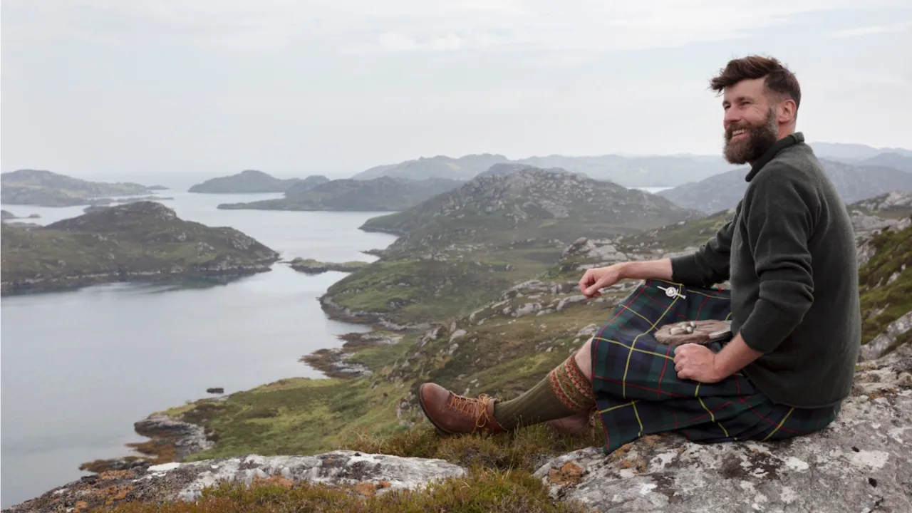 A smiling man wearing a kilt sits atop a cliff, water interspersed with rugged hills and islets below him