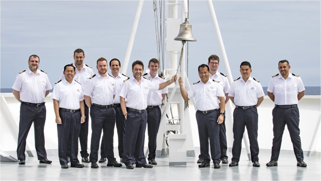 Quase uma dúzia de oficiais de navio, vestindo seus uniformes brancos e pretos, tocam um sino instalado em um navio