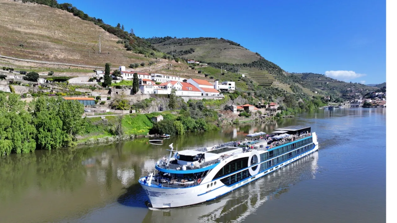 A long white river cruise vessel sails past a lush green riverbank filled with trees and houses