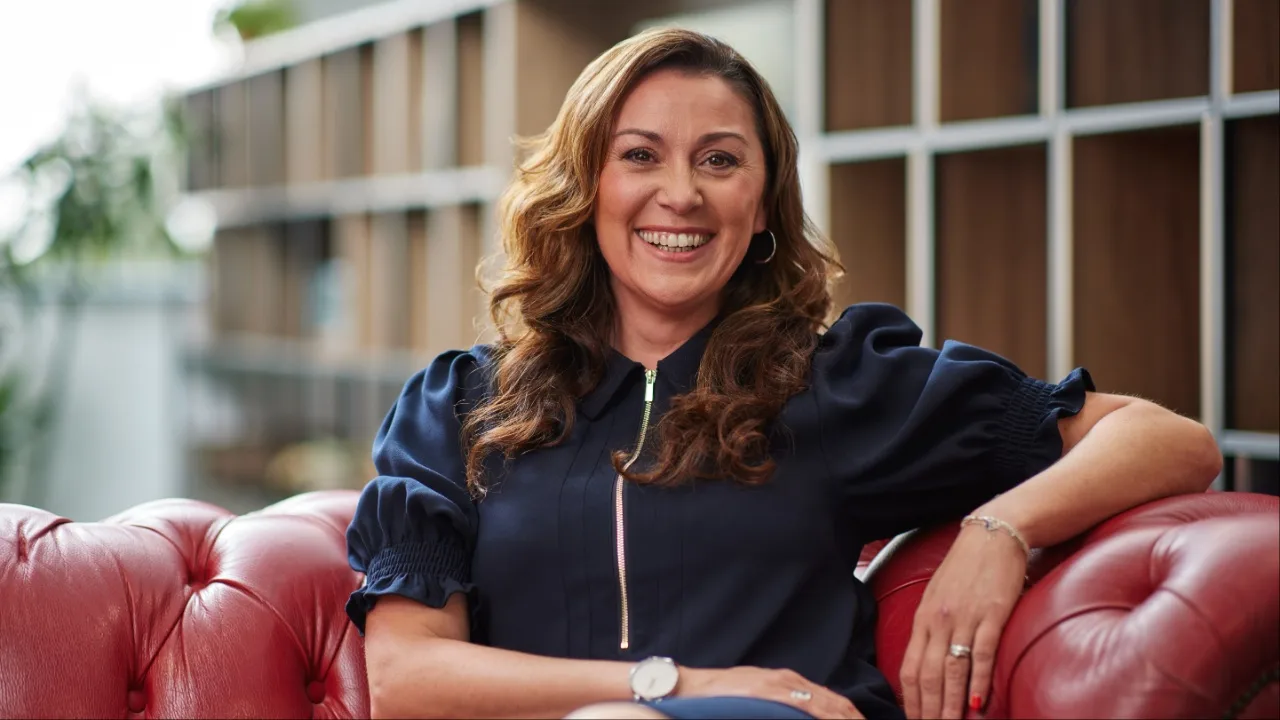 A smiling woman with thick, curly brunette hair, wearing a navy blue shirt, sat on a red leather sofa