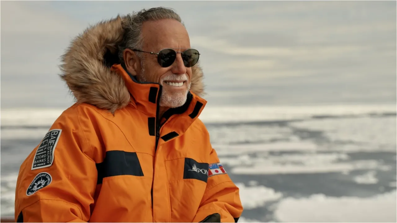 A smiling man with grey hair and dark sunglasses stares out over icy waters wearing a thick orange jacket